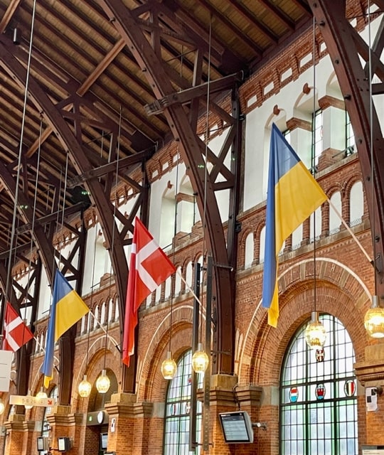 Inside Copenhagen Central Station.  Image shows tall arched windows with some stained glass and the flags of Sweden and Denmark.  The inside is smooth and flat and easy for people with all forms of disability, including wheelchair users to navigate around
