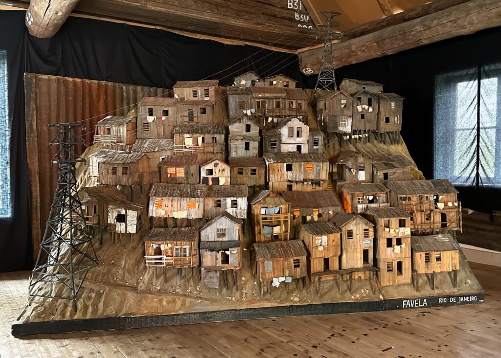 A sculpture of wooden houses on the side of a hill to replicate the favelas of Rio de Janeiro.  The sculpture is houses in building with wooden beams and is next to a small window.