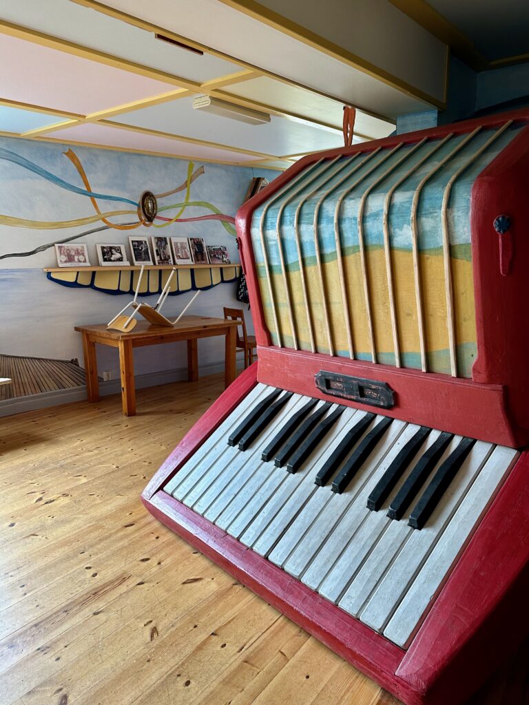 A sculpture of a giant wooden piano in red with a picture of a beach on the top of it. In  the background is a shelf with black and white pictures on it.