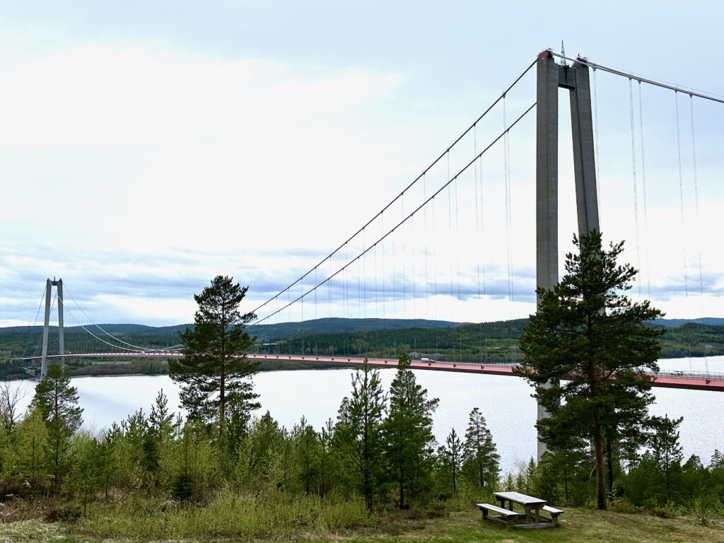 The High Coast Bridge, a large suspension bridge over the river with trees in the foreground and a picnic bench.  