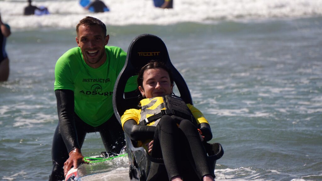 Molly is sitting on the adaptive surfing board and smiling while Cyril is behind her, guiding the board and laughing