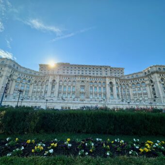 Picture shows a picture of the Palace of Parliament from the garden outside. The sun is shining and the sky is blue.