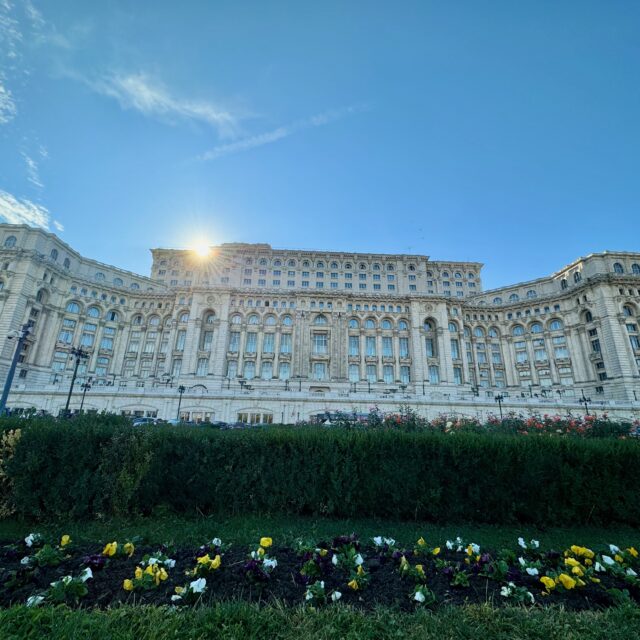 Picture shows a picture of the Palace of Parliament from the garden outside. The sun is shining and the sky is blue.