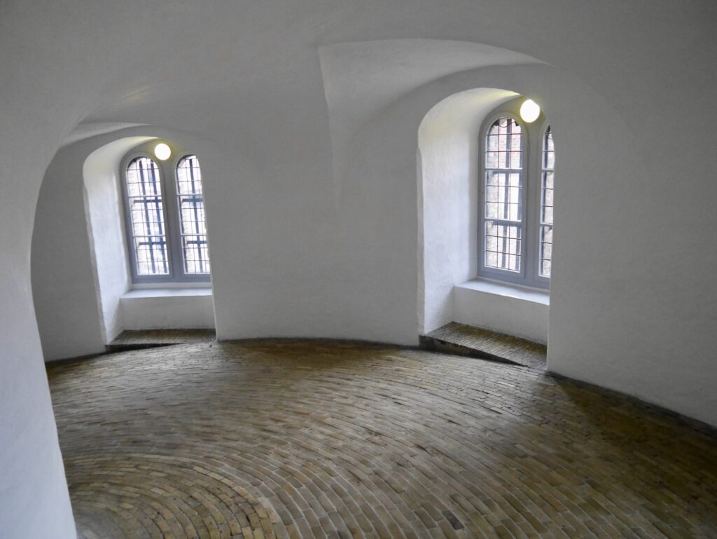 Image shows a wide and sloping path made of bricks laid end to end.  There are two large domed windows in alcoves with lights suspended above.  The walls are painted white.