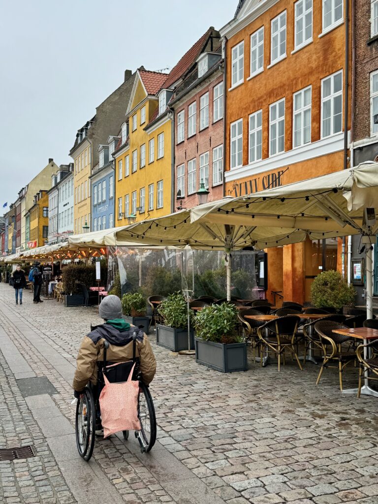 Image shows a young man in a wheelchair outside some of the restaurants lining the street.  He is wearing a brown coat and grey beanie and there is a light orange bag on the back of his chair.  The image also shows how accessible the are is with cobbles with smooth paths built in.
