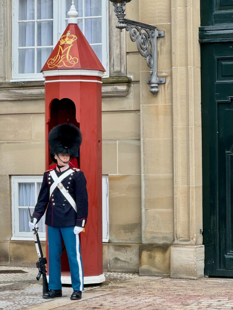 Image shows a soldier standing guard outside a red sentry box.  He wears a navy blue military jacket and mid blue trousers with white stripes down the legs.  Over his chest is a white cross belt.  On his head he wears a black bearskin hat.  He is holding a rifle and is standing to attention.  