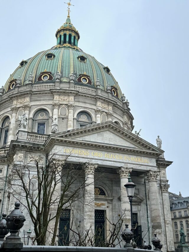 Image shows the external facade of a church. It is grey with an impressive domed roof in green and gold. The entrance has four large columns, above wihichis the inscription 'Herrens Ord Bliver Evindelig'