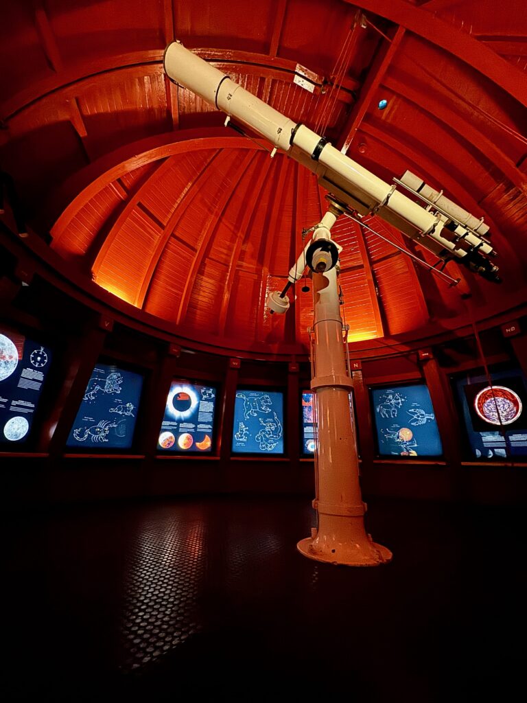 Image shows a large telescope in a circular room.  The domed ceiling is red and on the wall are many pictures related to the stars.