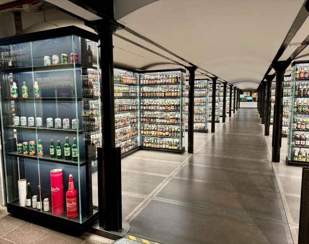 The image shows the bottle room at the House of Carlsberg.  There are many glass display cabinets showcasing the hundreds of different beers produced throughout the world.  These are displayed in aisles with a wide walkway running down the centre of each.
