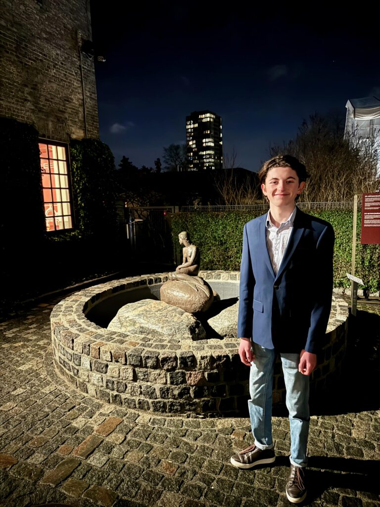Photo is taken at night.  In the foreground is a teenage boy, smartly dressed in a navy blazer, white shirt and jeans.  He is smiling and standing next to a small stone statue of a mermaid on a rock.  the statue is in the middle of a circular fountain in a cobbled accessible courtyard.  In the background is a tall illuminated building.