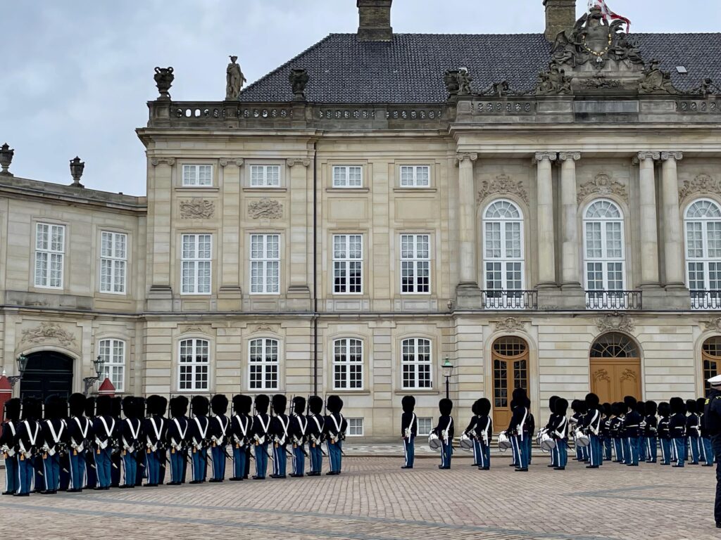 Many soldiers are stood in rows on a large paved square outside am impressive beige stone building.  They are dressed in the uniform of the Danish army and many are holding instruments.