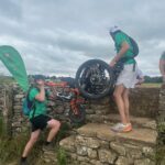 Tow men wearing green t-shirts are lifting a wheelchair over a stile in a dry stone wall in the British countryside.