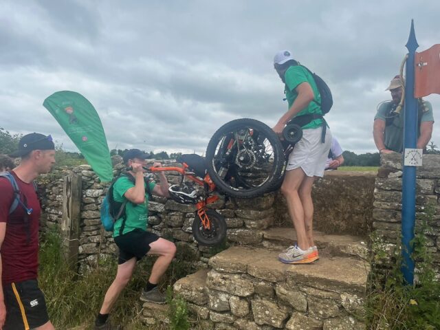 Tow men wearing green t-shirts are lifting a wheelchair over a stile in a dry stone wall in the British countryside.