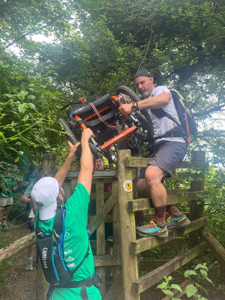 A group of men are passing an all terrain wheelchair over a kissing gate in the countryside.  One is standing on the gate itself while another is passing it up to him.