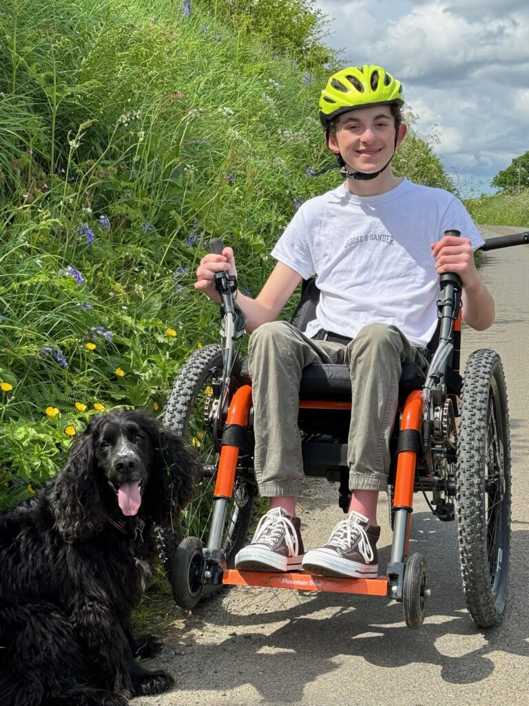 A young man is sitting in a bright orange wheelchair in a Devon country lane.  he is smiling and wearing a white shirt and khaki combat pants.  His dog, a black cocker spaniel is sitting next to him, panting,