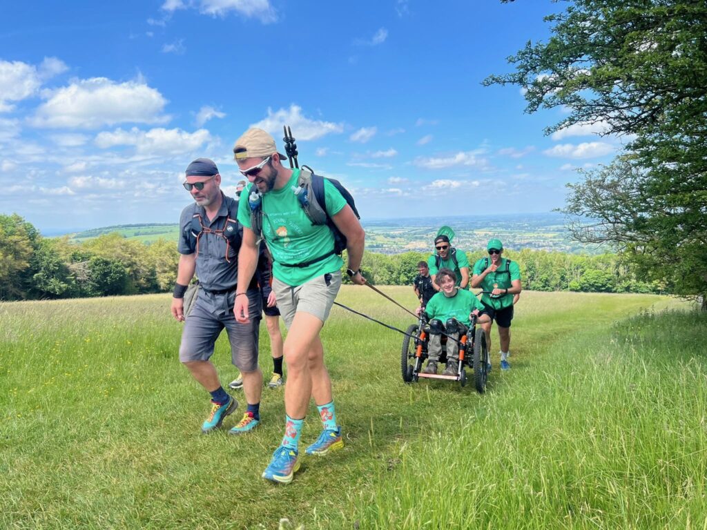 A team of men are pulling and pushing a young man in a wheelchair through a field.  the filed has a slight uphill incline.