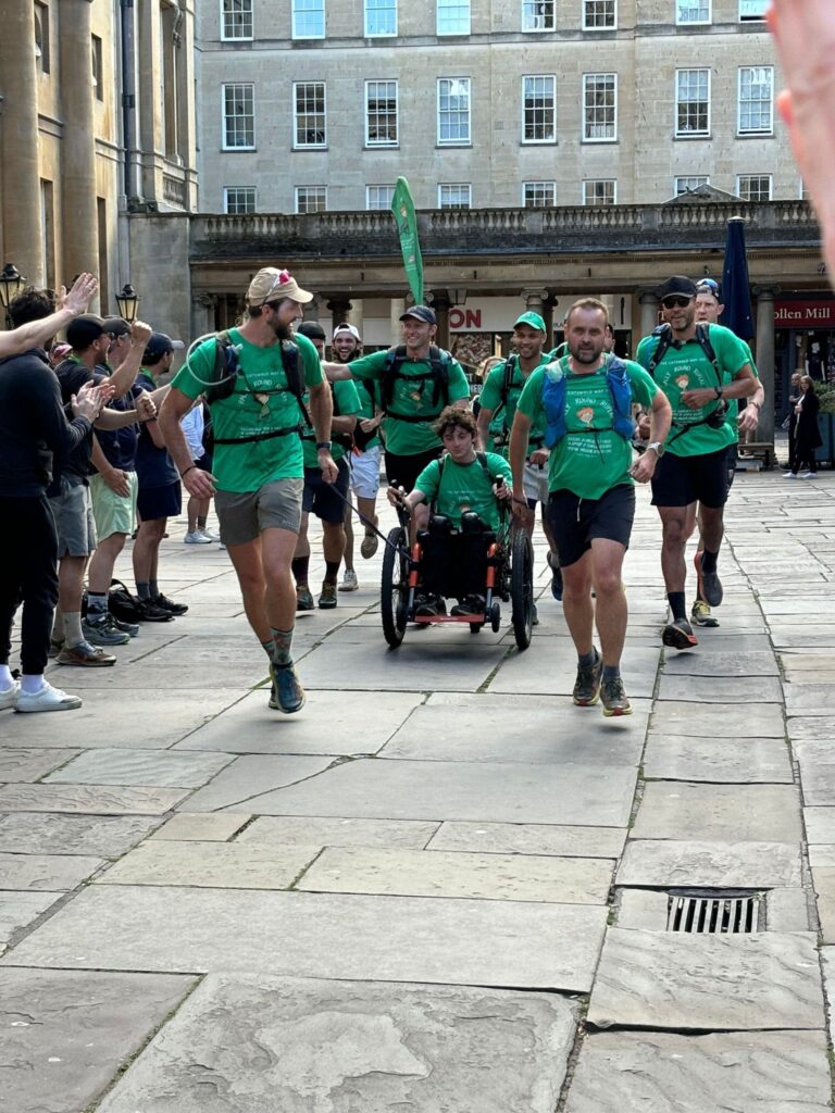 A small team, all wearing green Rally Round Rupert t-shirts are running through the street near Bath Abbey.  They are pushing and pulling a young man in a wheelchair who is also helping to propel it.  All look tired but happy and triumphant.