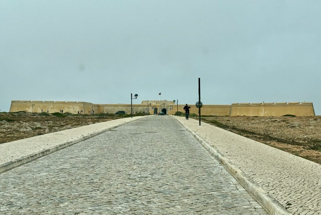 The fortress at Sagres.  It is built of yellowish stone and dominates the landscape.  A wide road approaches it.