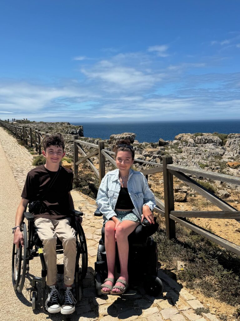 A young mana nd a young woman are in wheelchairs on a wide path with views out to sea.  Both are smiling and the sun is shining brightly.