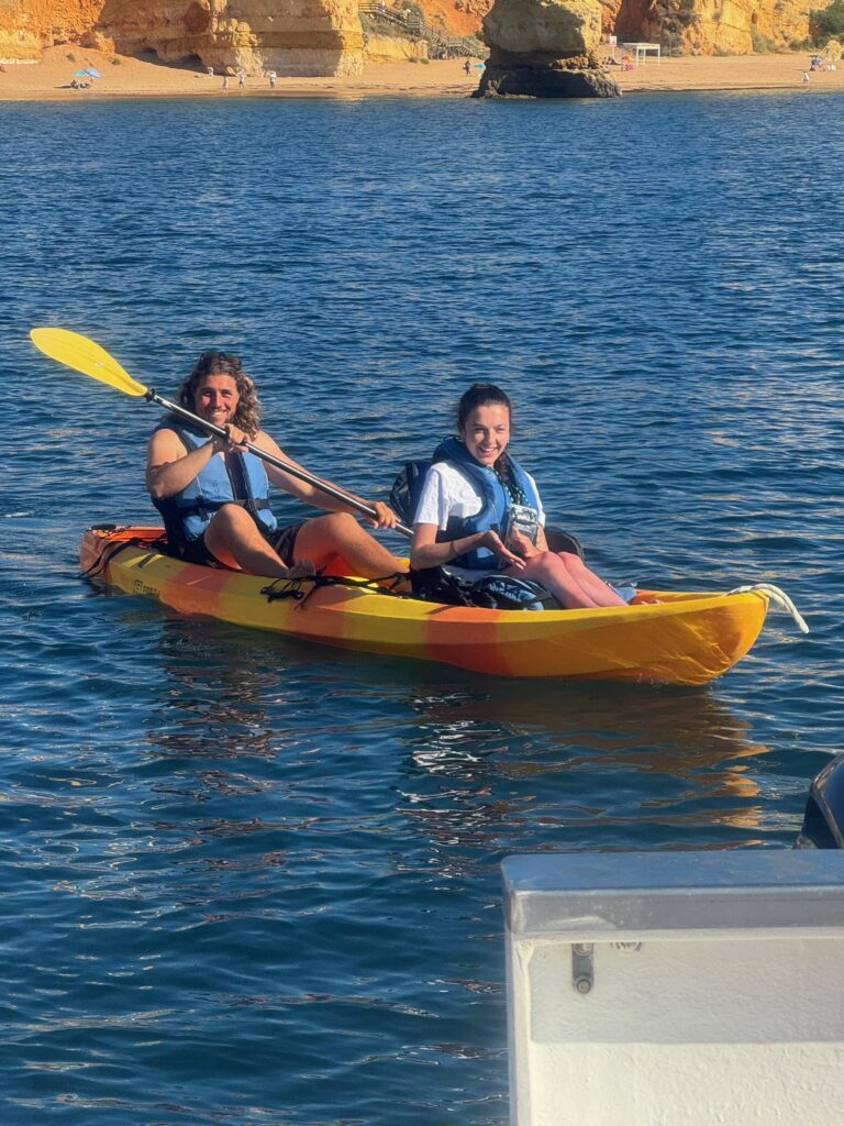 Two people are sitting in a kayak.  A man is behind and is holding a paddle aloft.  The young woman in front is smiling broadly.  Both are waring blue life jackets.