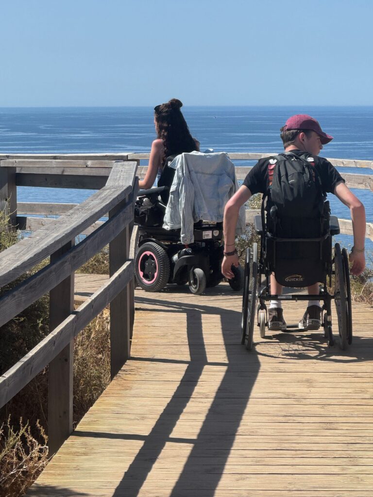 Two young people in wheelchairs are moving along wooden boardwalks looking out to sea.