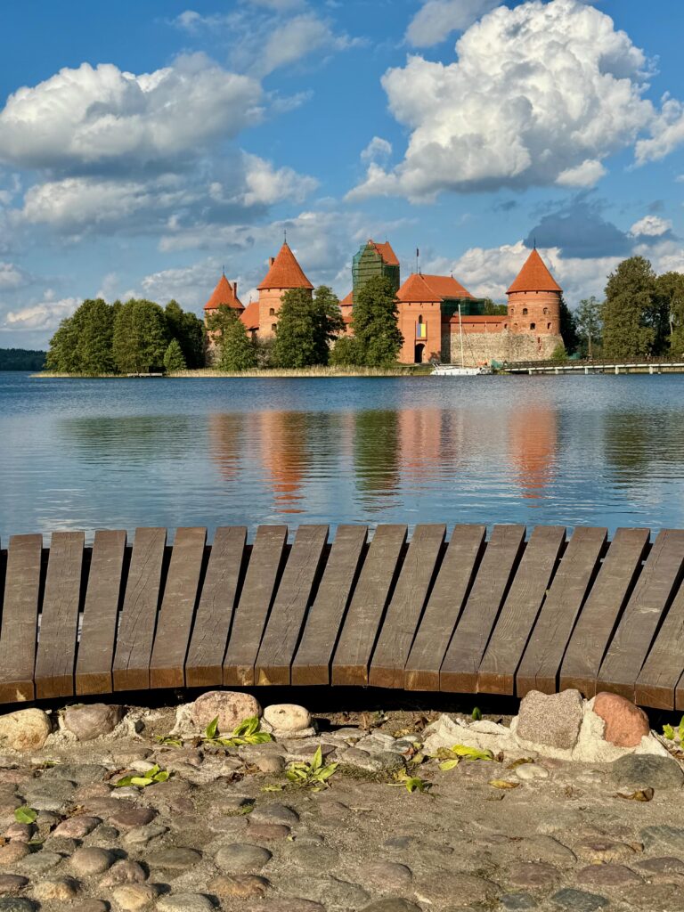 A fairytale type castle surrounded by trees is on an island on the far side of a lake.  There is a wooden walkway on the near side.  The sun is shining and the water is very blue.