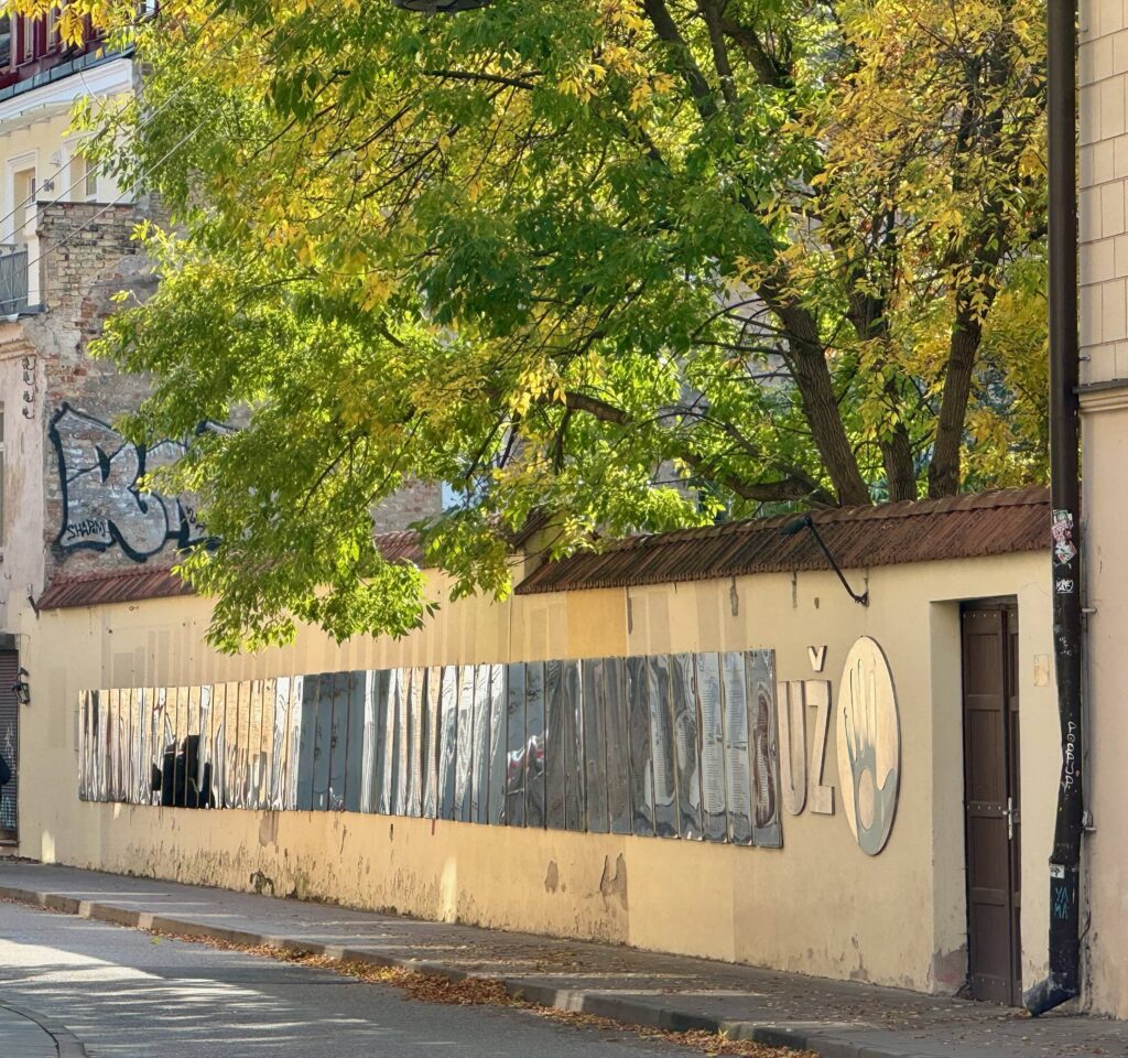 A yellow wall with metal plaques.  Each one is a copy of the constitution of Uzupis in 44 different languages.
