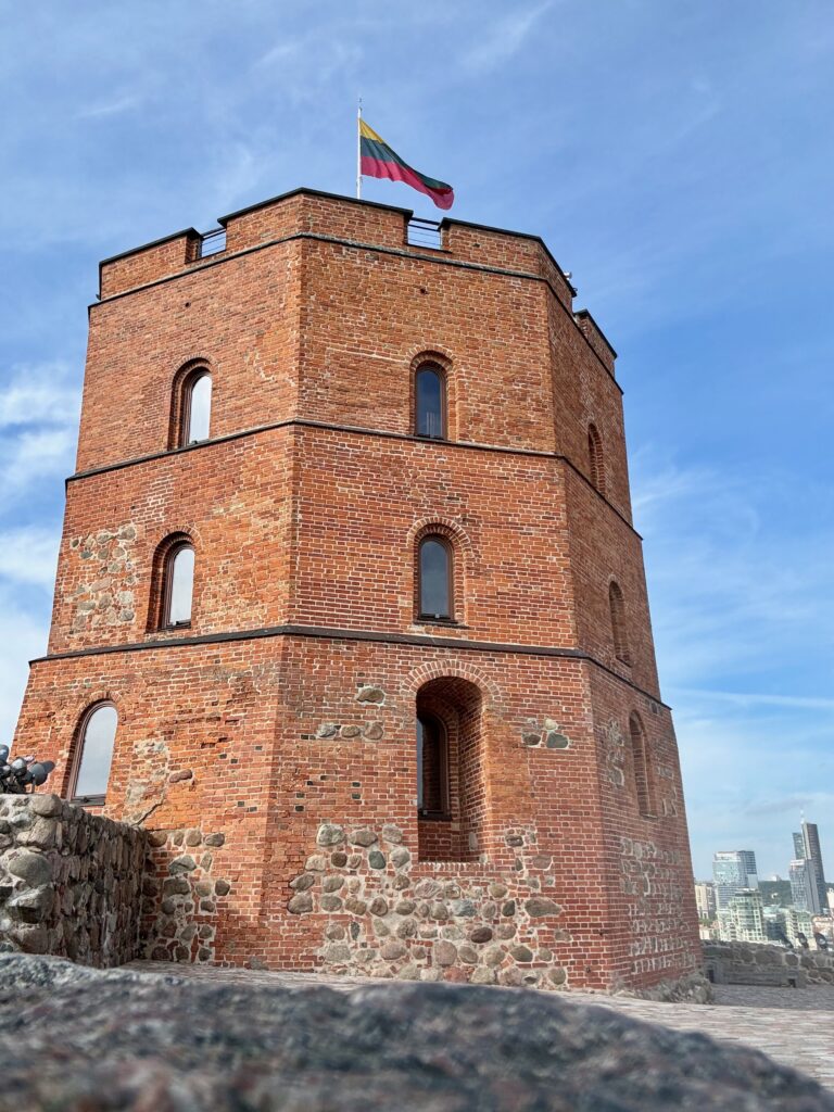 Image shows the Gendiminas Tower - it is a red brick building with a hexagonal structure.  The battlements at the top display the Lithuanian flag of yellow, green and red.  In the background is a view over Vilnius.