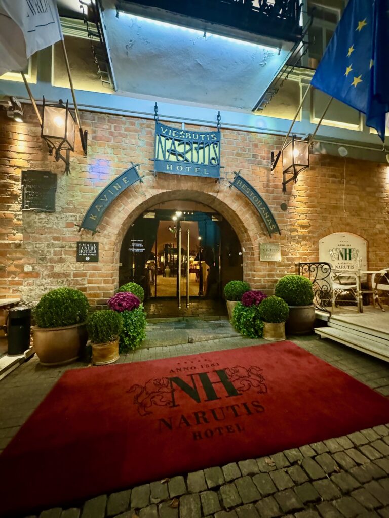 Exterior of the Hotel Narutis in the centre of Vilnius.  There is a brick wall with a large arched doorway.  Glass doors open onto a cosily lit inter.  Outside their a large red carpet flanked by pots containing green box balls.