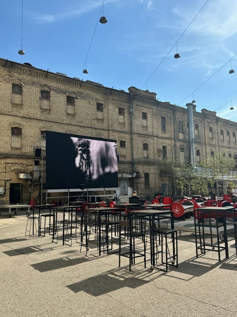 A prison yard in front of an old style Soviet prison.  There are tables and chairs in front along with a large screen displaying a picture of a woman.