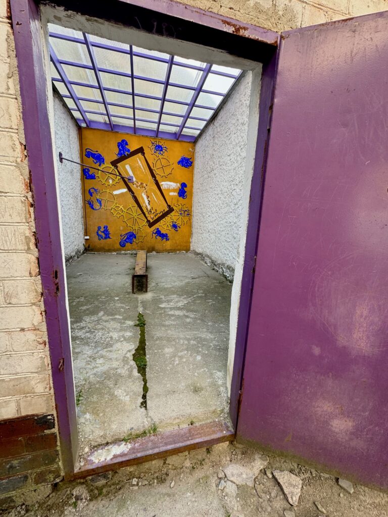 A windowless cell - there is a bench in the middle and a decorated panel at the back.  The roof is semi transparent and there is a purple door.  