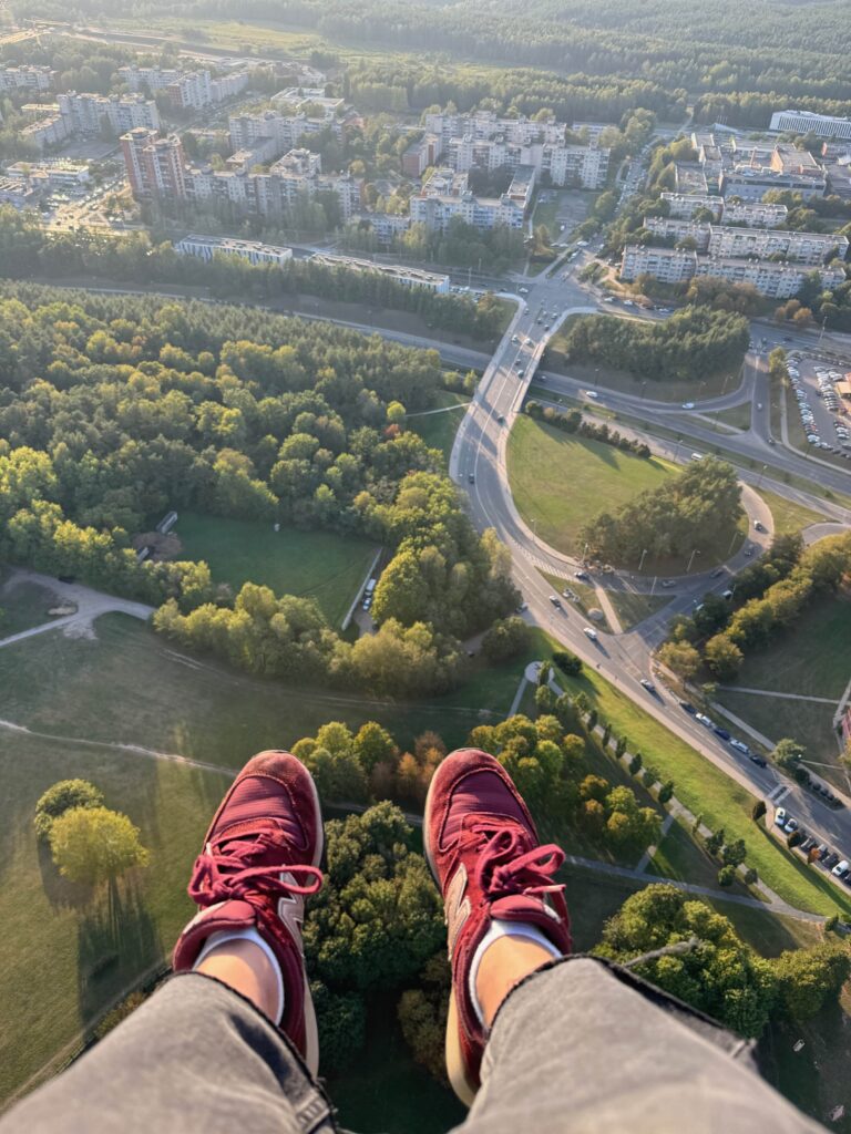 A pair of feet protrude over a ledge beneath which is a view of Vilnius from a long way up! Below there are tall buildings, a large road cutting through the middle and lots of greenery.