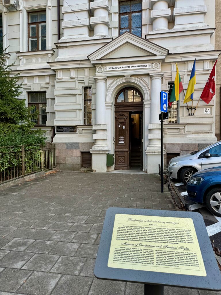 A white stone building with a large wooden door which is partially open.  Outside is a metal plaque identifying as the Museum of Occupation and Freedom Fights.