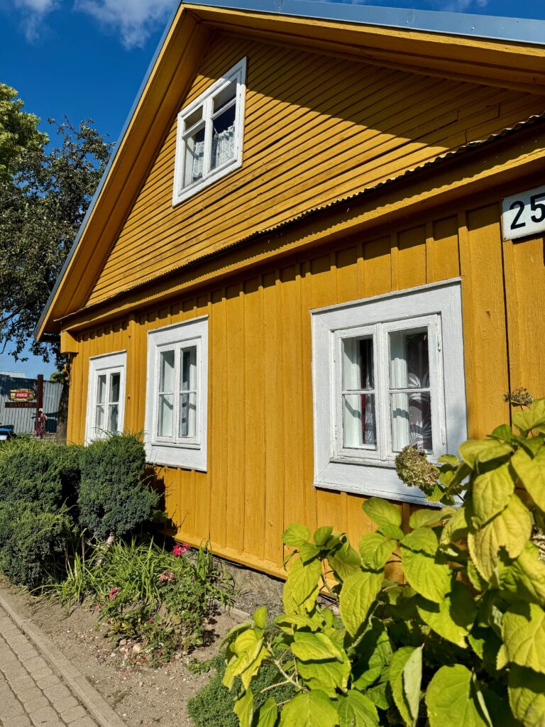 A mustard yellow wooden building - it has white windows and the number 25.  The sky is very blue and there are green bushes in front of the house.