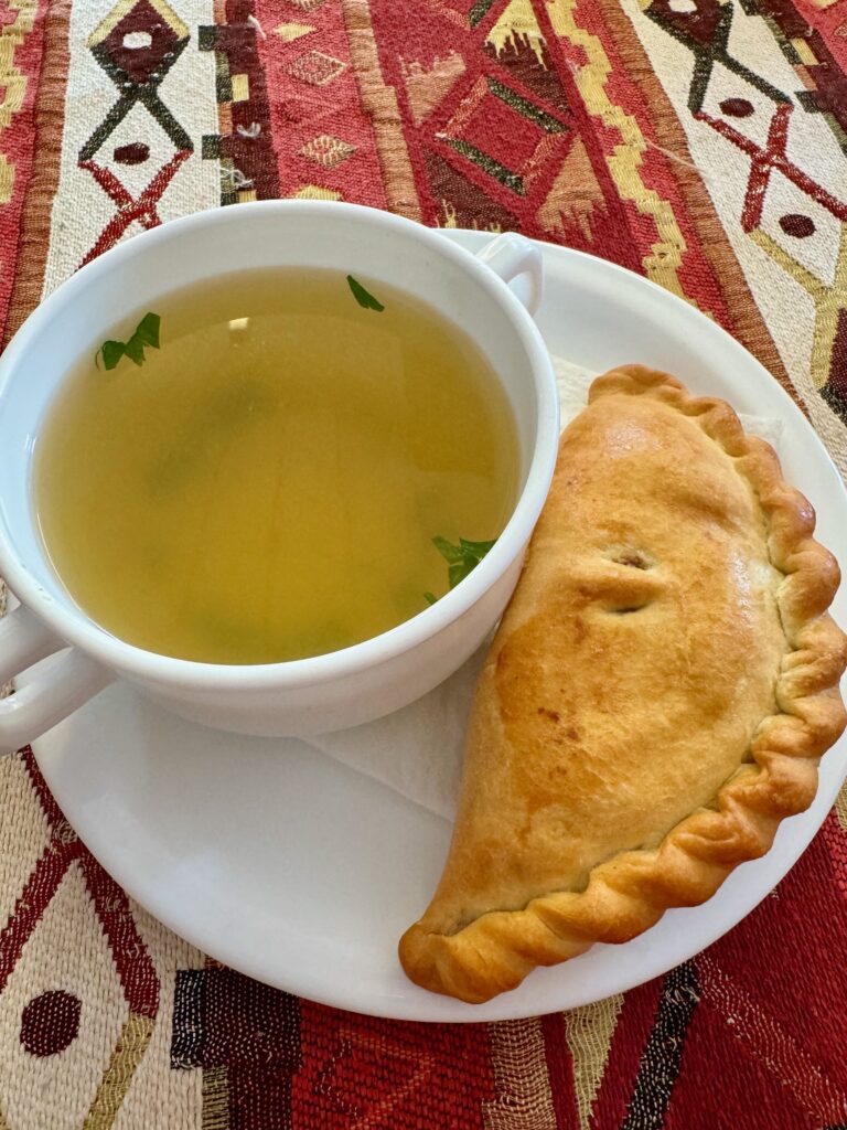 A cup of broth and a traditional kybyn on a white plate.  The plates on a mostly red patterned tablecloth.