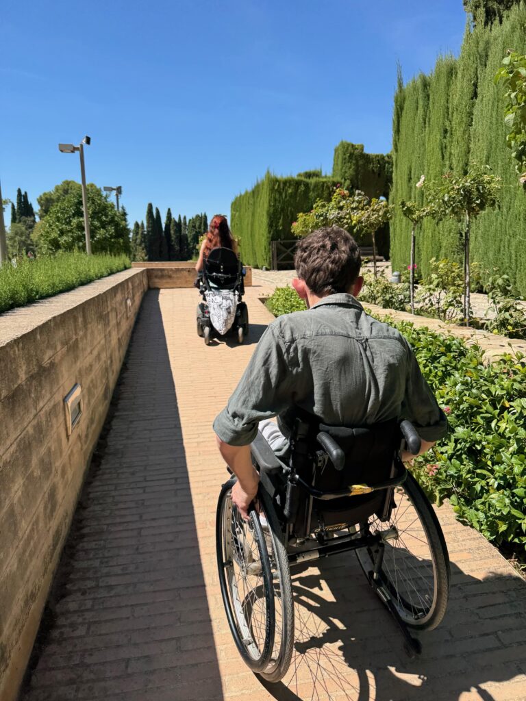 The image depicts two wheelchair users, one in a power chair, the other in a manual going up a smooth ramp to access the gardens of the Generalife at the Alhambra..