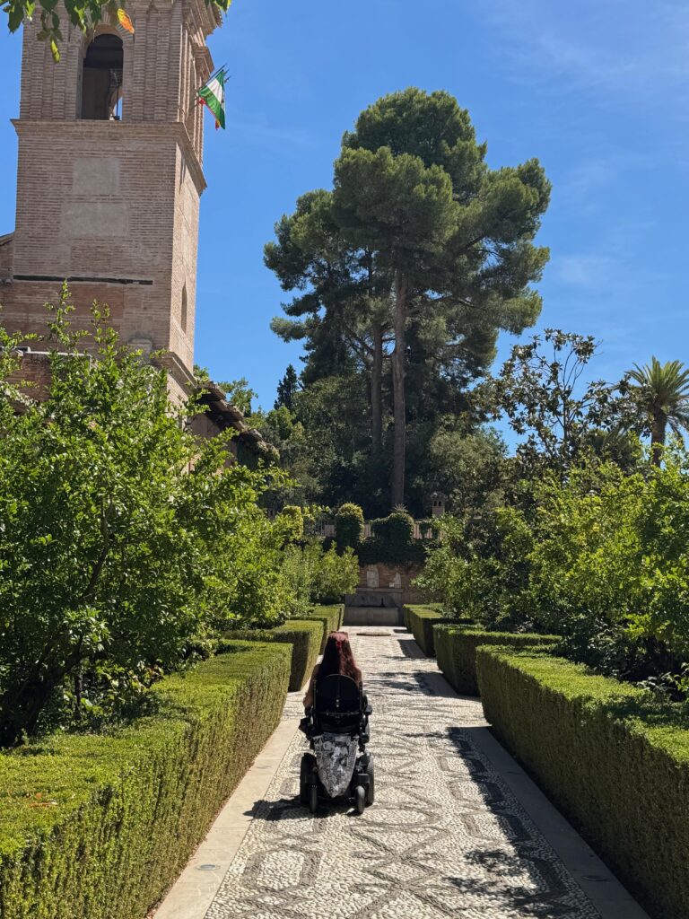 A gently cobbled path runs through the middle of the gardens of the old convent athlete Alhambra Palace.  A powerchair user is shown confidently driving along the path.