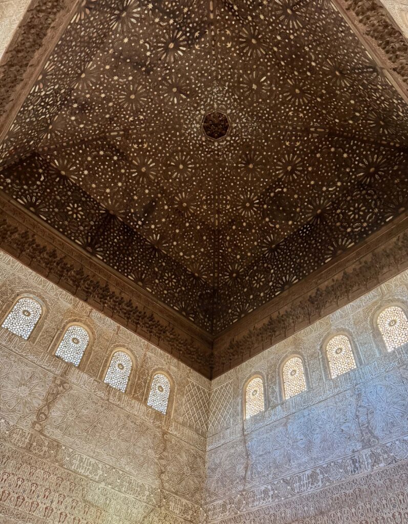 The image depicts a high ceilinged room with a domed wooden ceiling. This is intricately carved, covered with many stars. The walls are also embellished with carvings, with high, decorated windows running all the way around. 