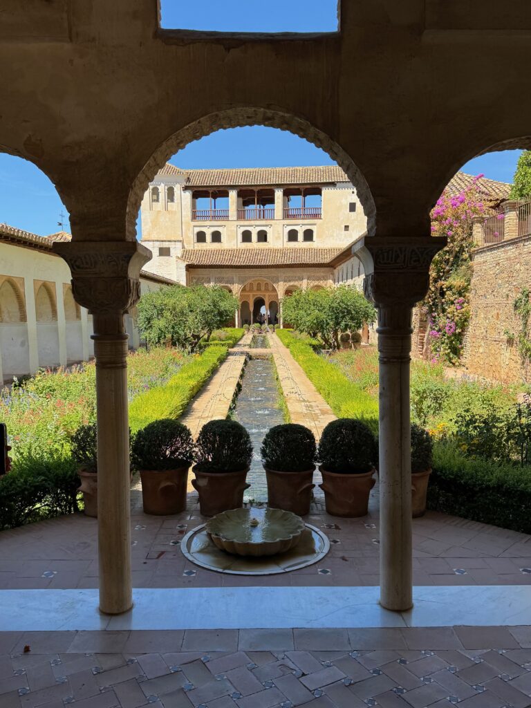 The image depicts the palace of the Generalife shown from the main entrance.  A central body of water, seen through a Moorish arch is framed on both sides by glorious gardens,
