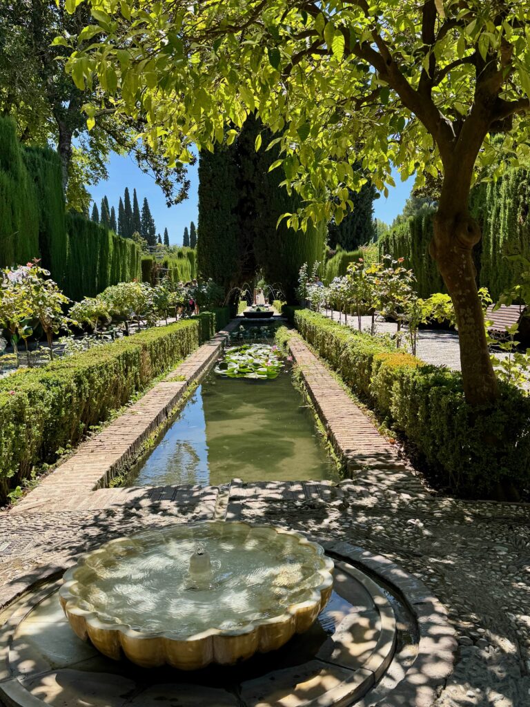 A wide rill is framed by a small border along with lush greenery.  Fountains are interspersed along the length of it.
