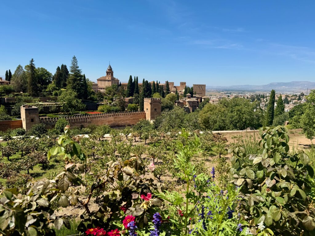 The foreground is filled with greenery and flowers while in the distance are the stone buildings of the Alhambra Palace. Beyond that lie the mountains of the Sierra Nevada.