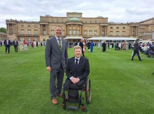 Picture shows two men at the King's Garden Party, on the lawn with Buckingham Palace in the background. One is standing, dressed in a grey suit with brown shoes and tie made of Cornish tartan. The other is seated in a wheelchair, dressed in a dark suit and dark tie. Both have big smiles on their faces. 