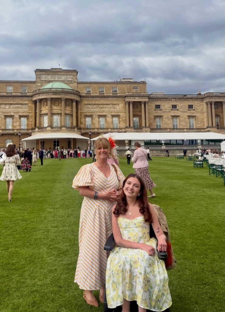 The phot shows two women on the lawn with Buckingham Palace in the background, waiting to meet the King. . One is a young woman with long dark heart in a yellow floral dress. she is in a wheelchair and is next to a blonde woman in an orange and white striped dres. She has an orange fascinator in her hair.Both look very happy, with big smiles on their faces.